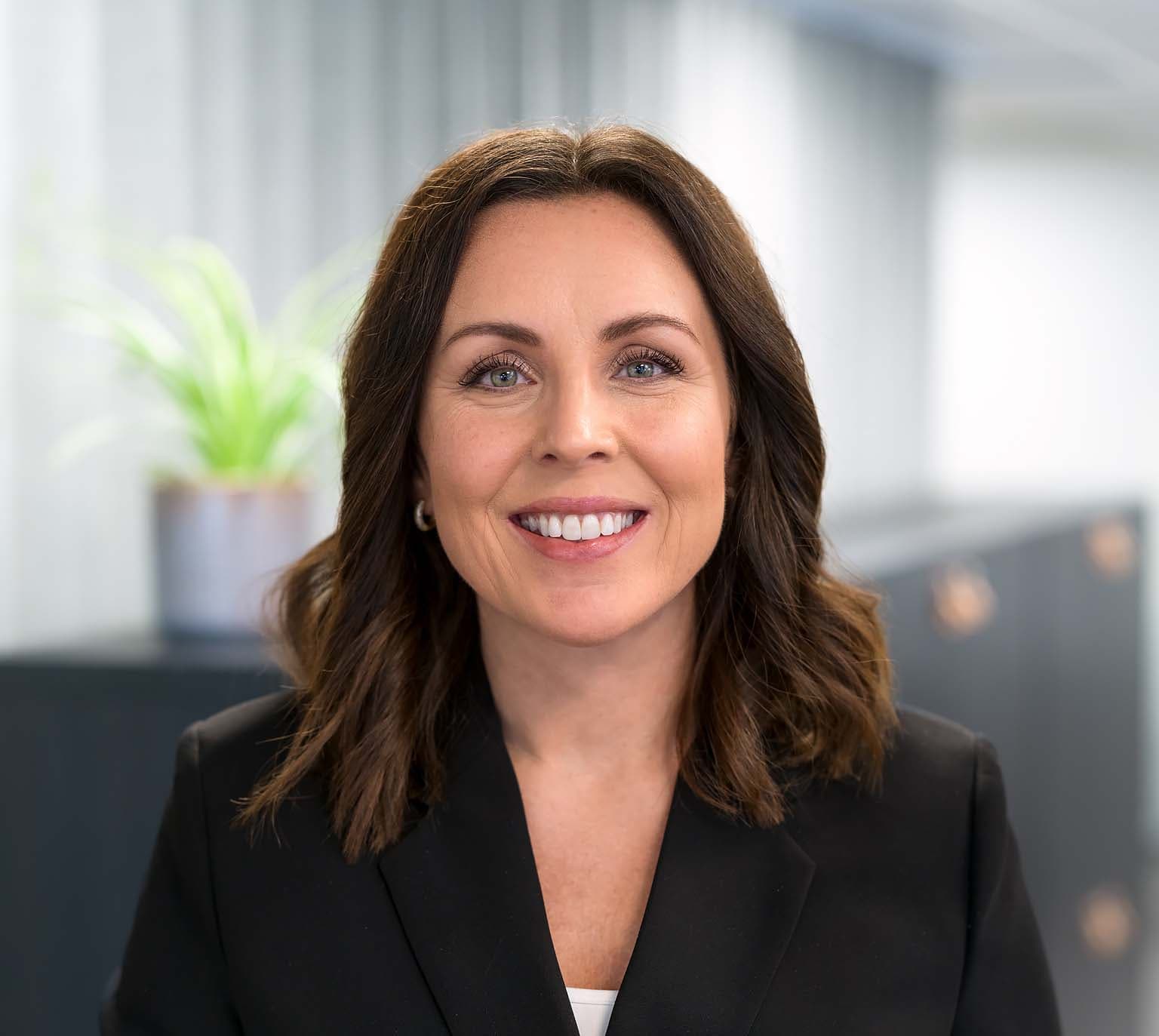 Smiling woman with brown hair in a black blazer, standing indoors with a blurred plant in the background.