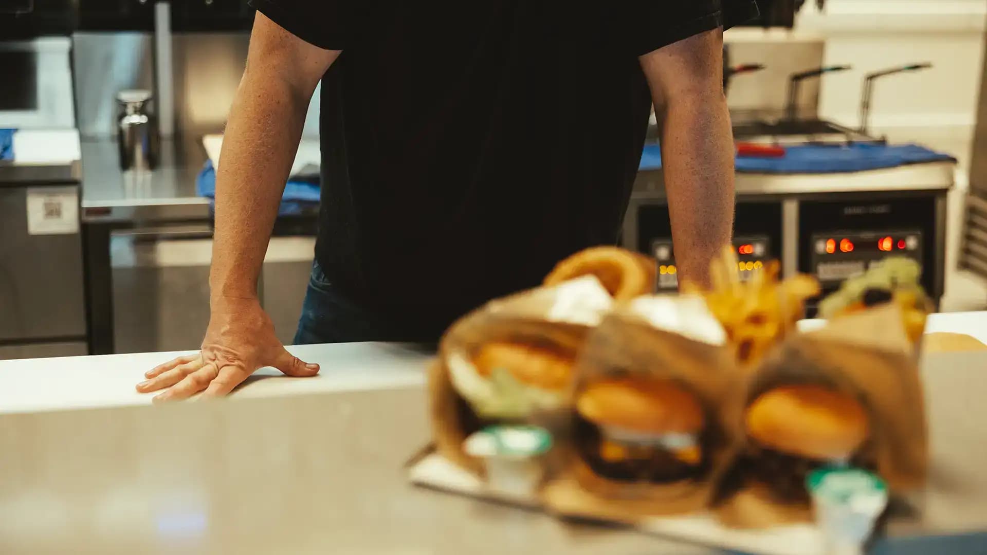 En plåt med paketerade hamburgare i förgrunden, bakom står en man och lutar sig framåt mot köksbänken.