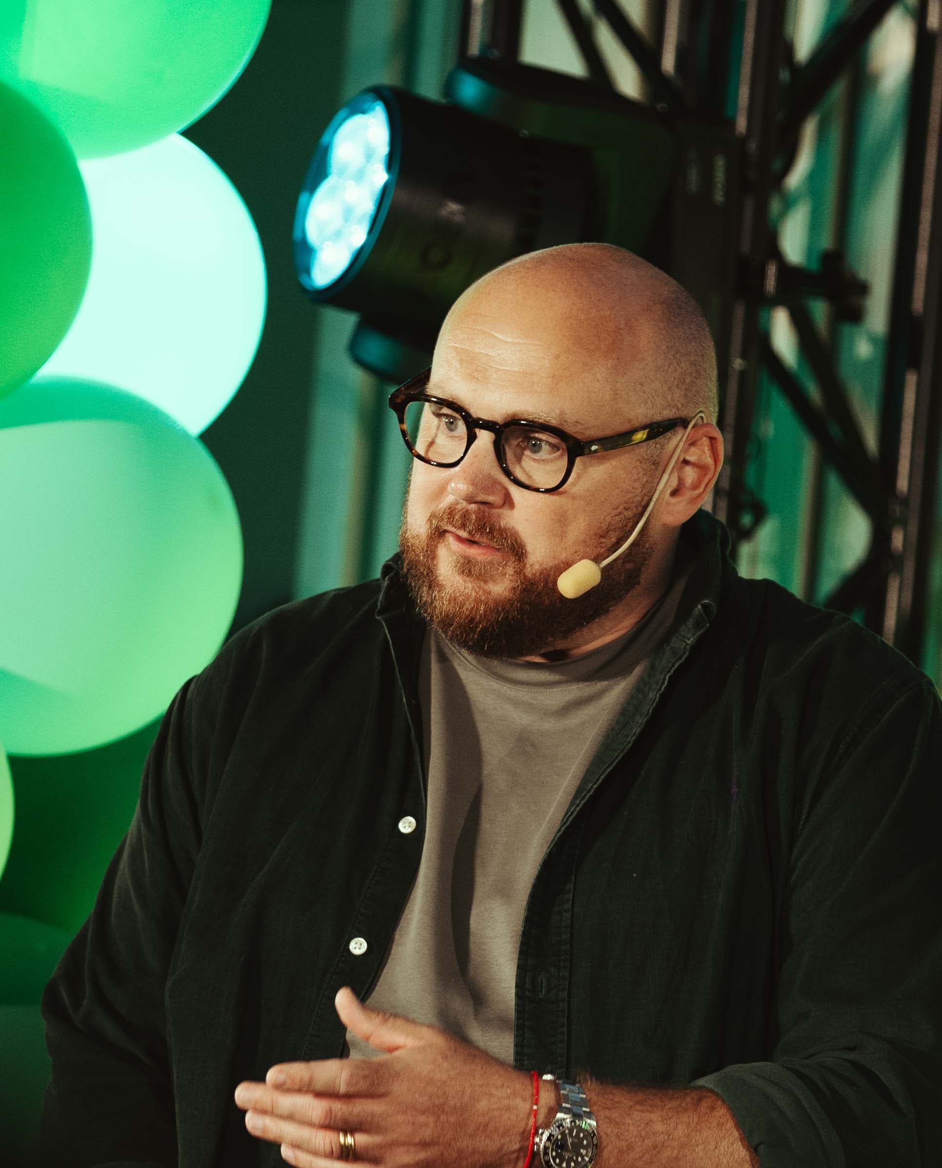 A man wearing glasses and a headset mic speaks on stage, with green balloons and stage lighting in the background.