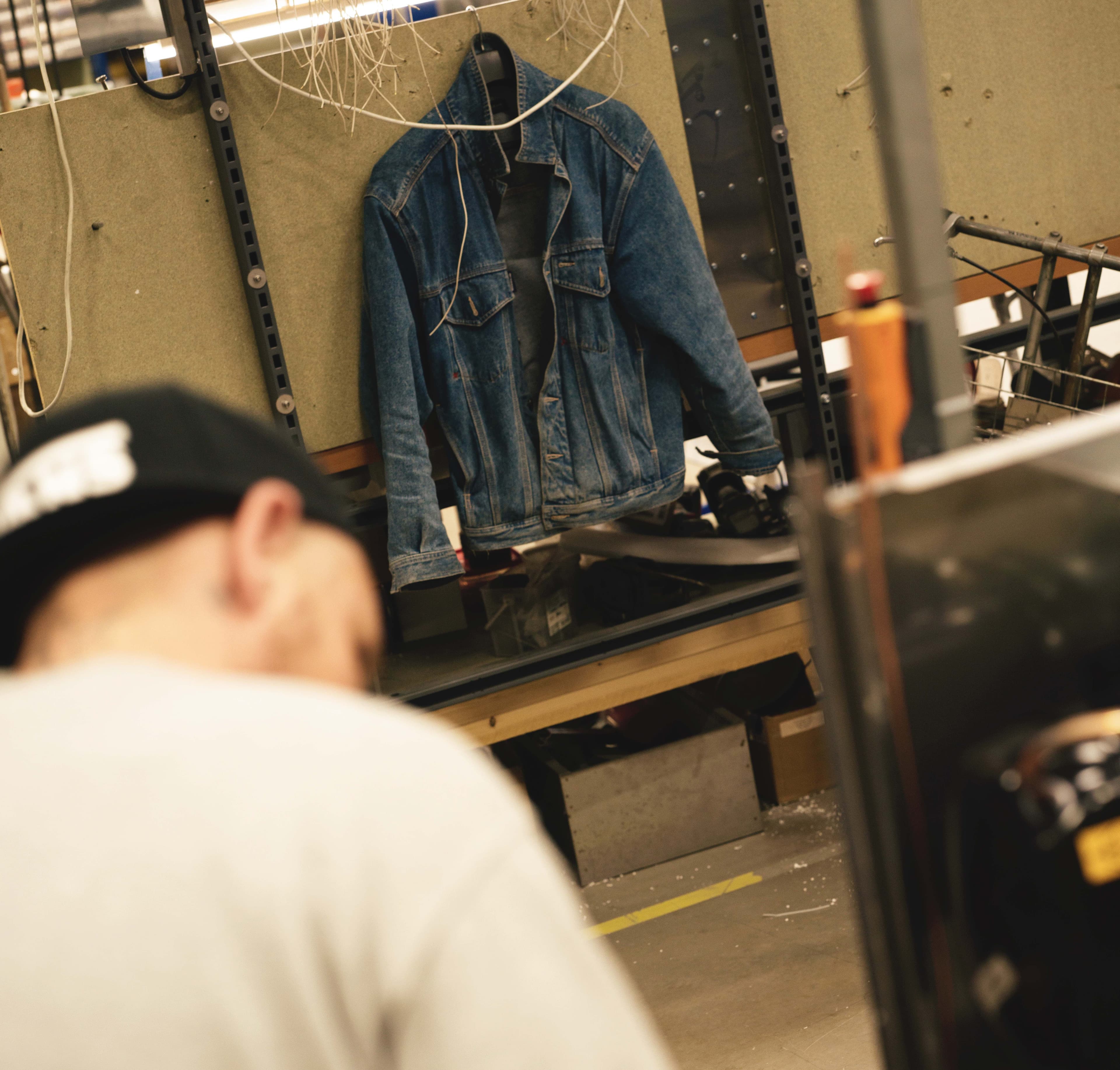 A denim jacket hangs on a pegboard in a workshop. A person with a baseball cap is blurred in the foreground, facing away.