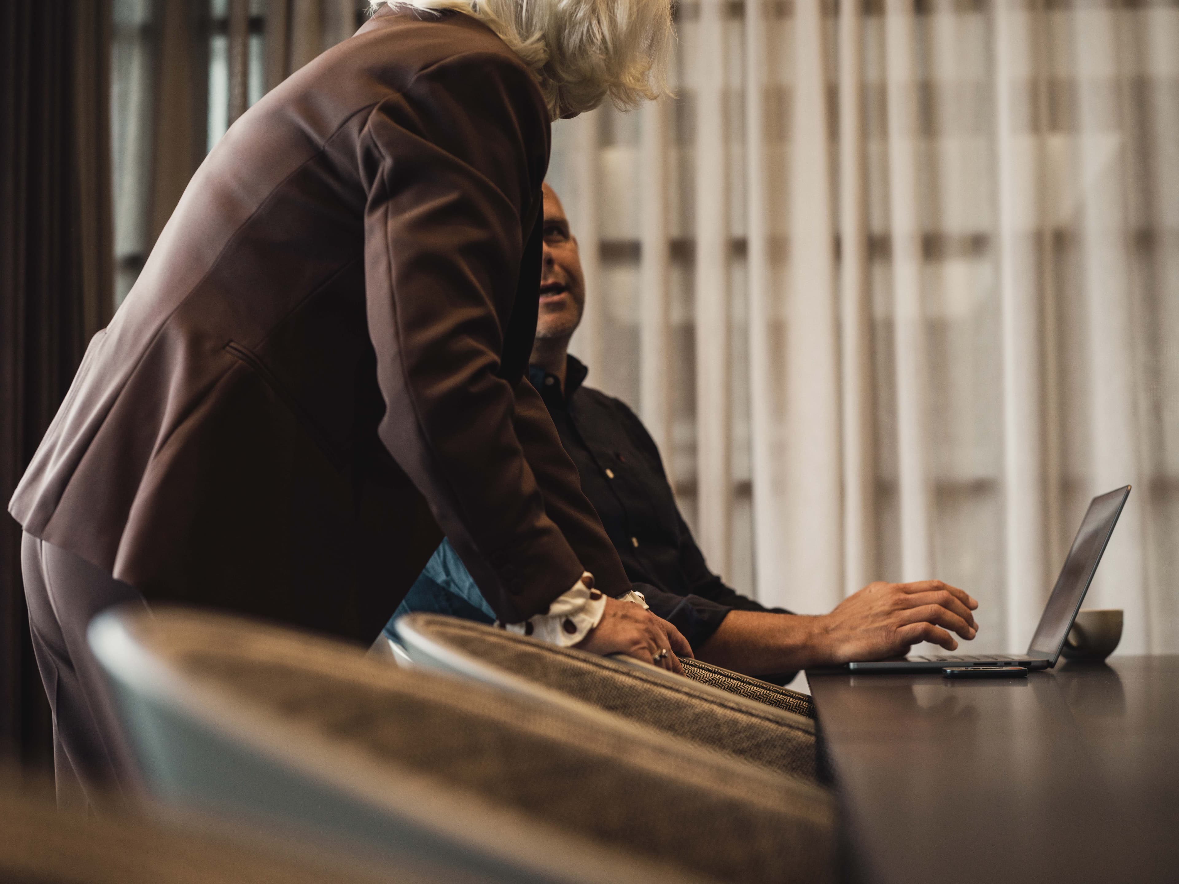 Two people engaged in conversation; one is seated at a table with a laptop while the other leans in, suggesting a collaborative discussion.