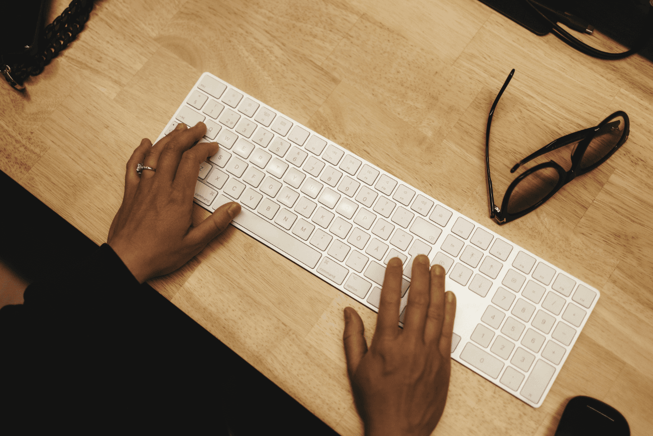 Hands typing on a white keyboard on a wooden desk, with glasses placed nearby.