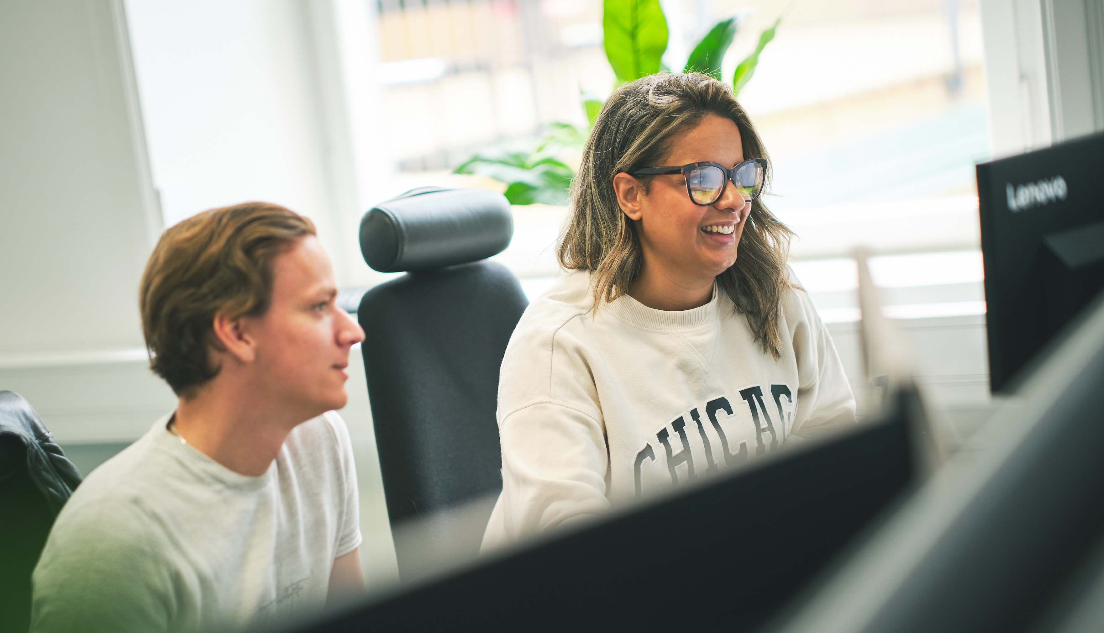 Two people working together at a desk with computers, smiling and looking at a screen, with plants in the background.