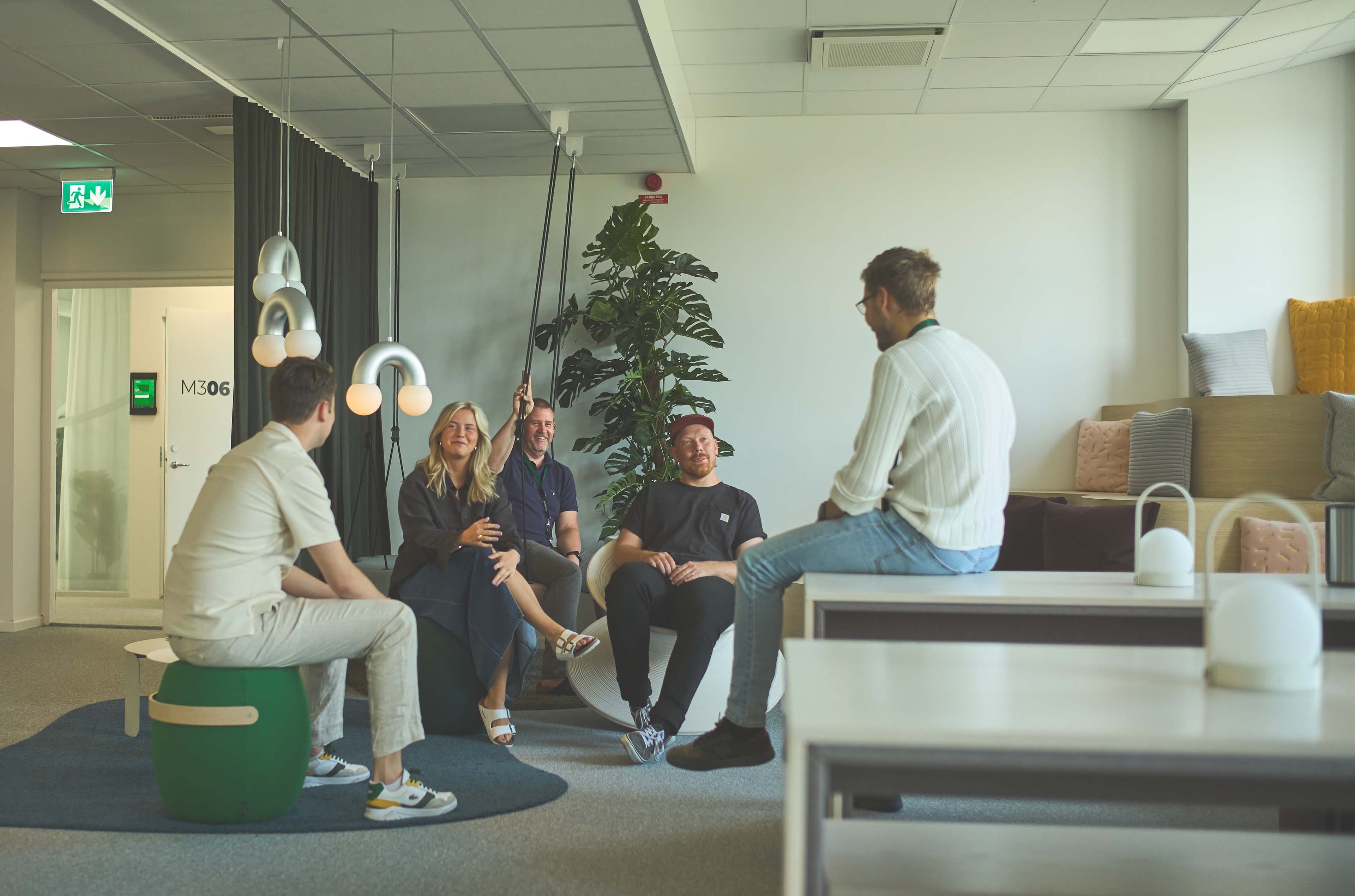 A group of five people sitting and talking in a modern, casual office space with plants and stylish decor.