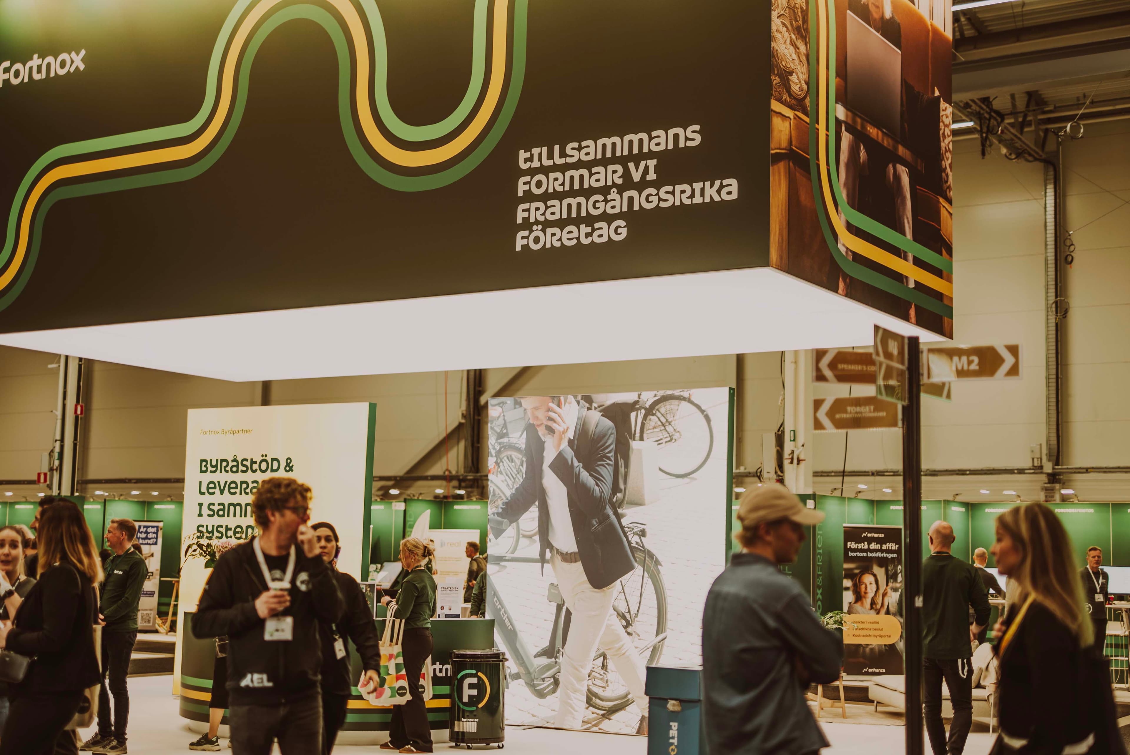 Trade show booth with people walking around, large signage overhead, and a poster of a person using a phone and bicycle in the background.