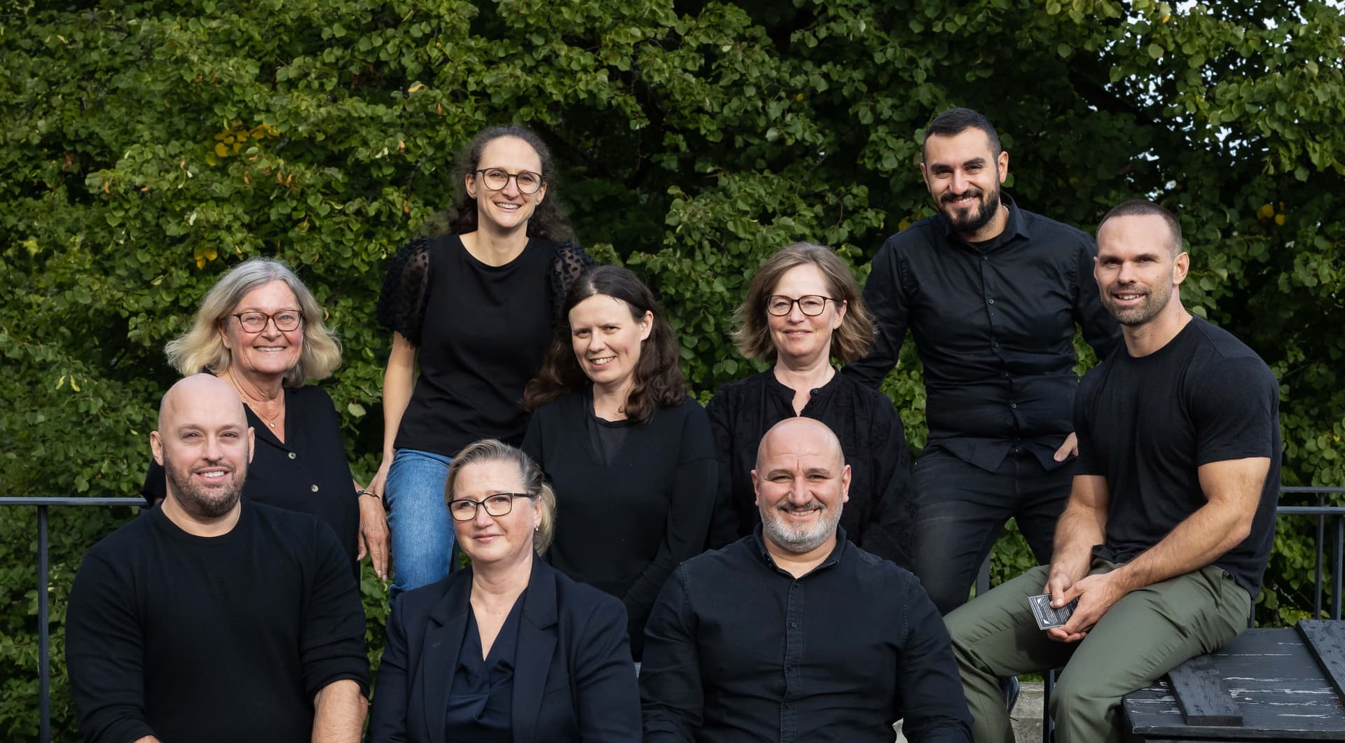 Group of ten people smiling, dressed in black, sitting and standing outdoors with lush green foliage in the background.