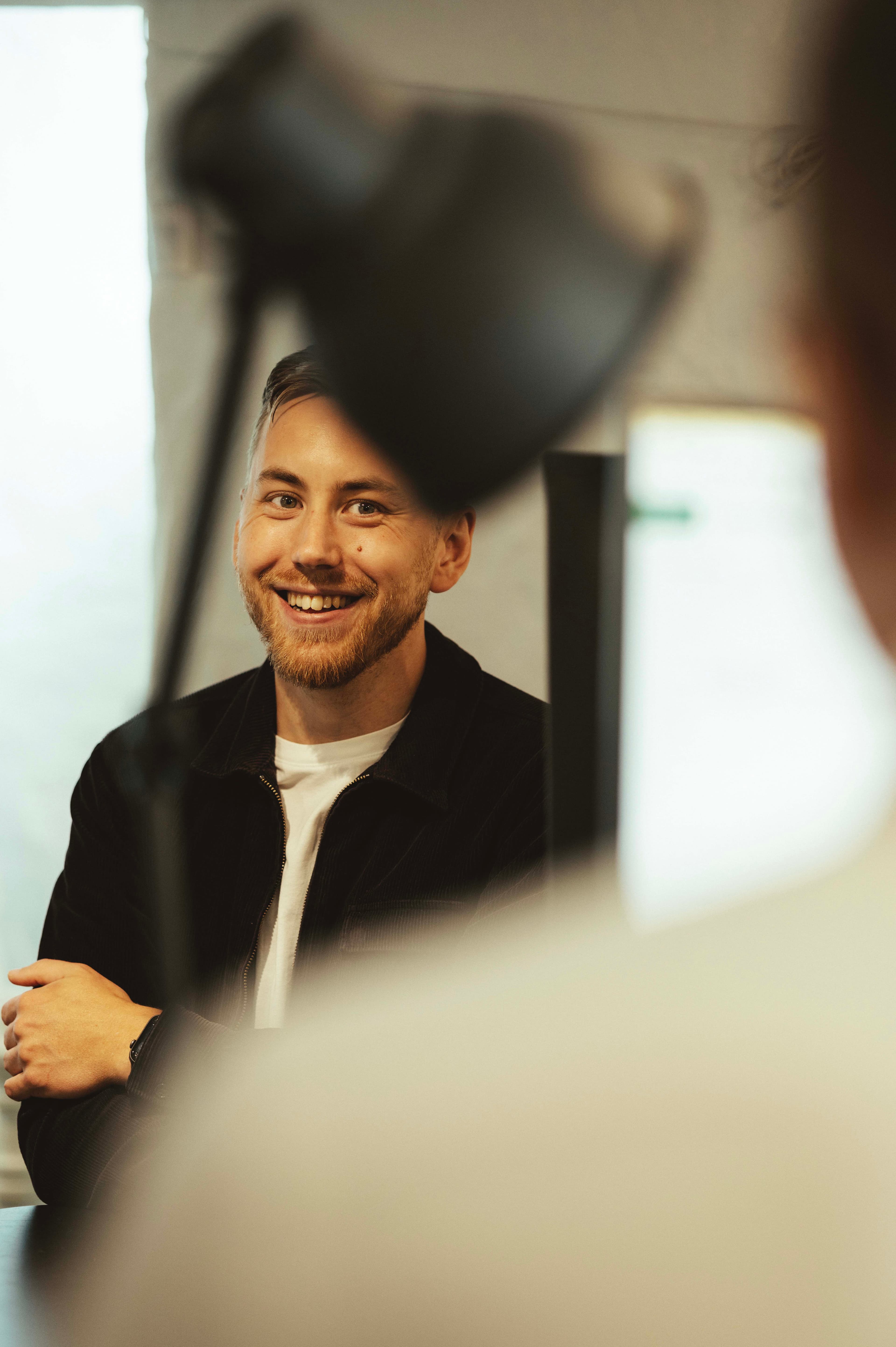 Smiling man in a black jacket and white shirt, partially obscured view, sitting indoors with a blurred foreground.