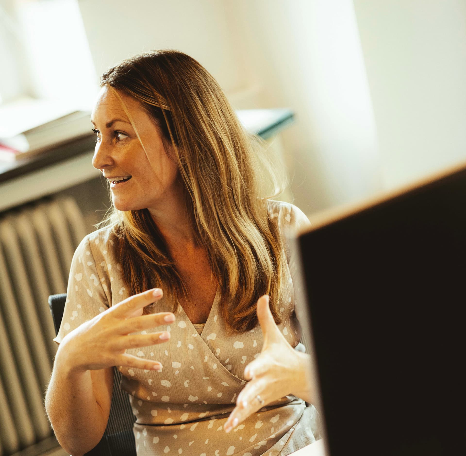 Woman with long hair gesturing while talking, seated indoors near a window with soft lighting and part of a computer screen visible in the foreground.
