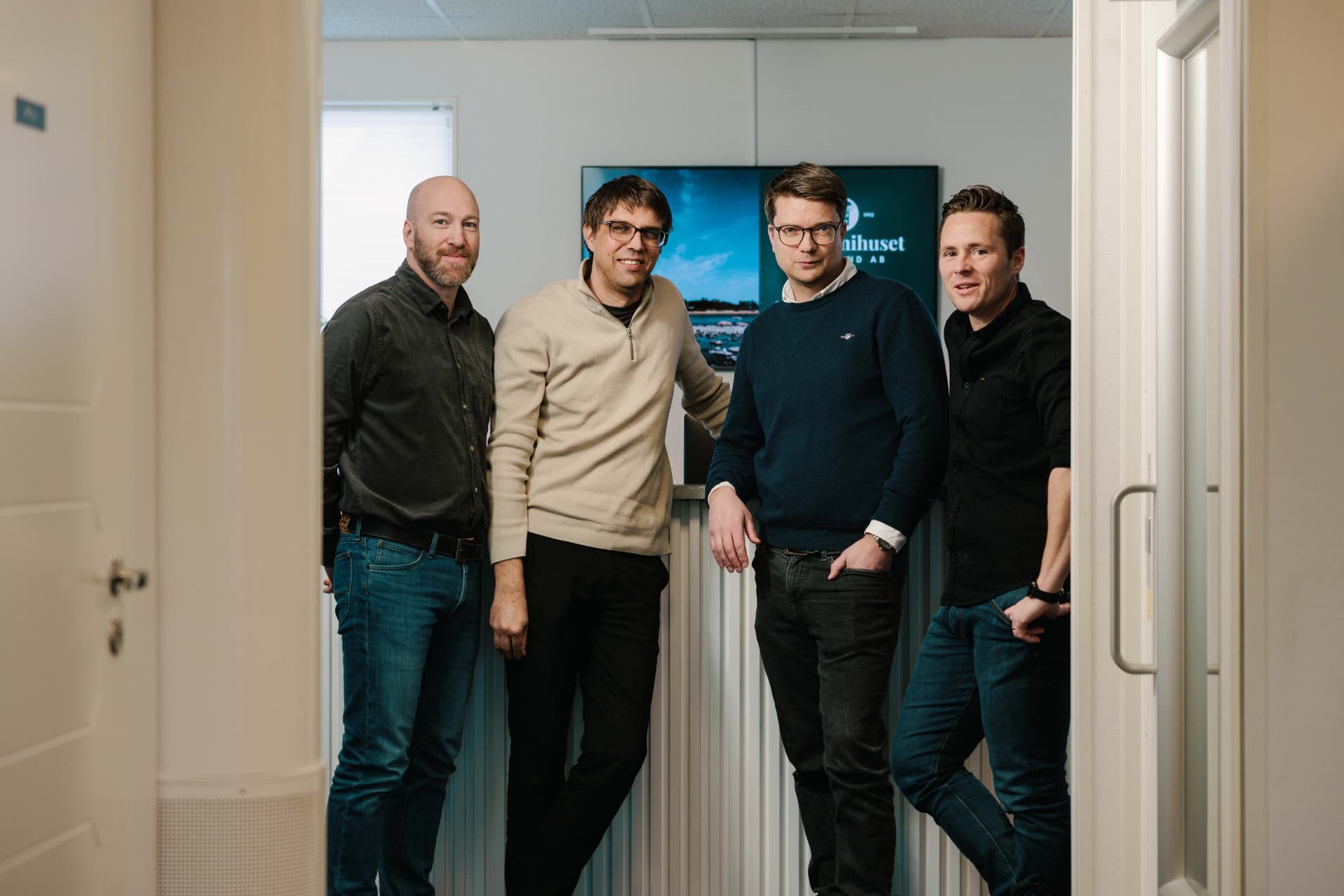 Four men stand together in a hallway, casually dressed, smiling at the camera with a sign in the background.