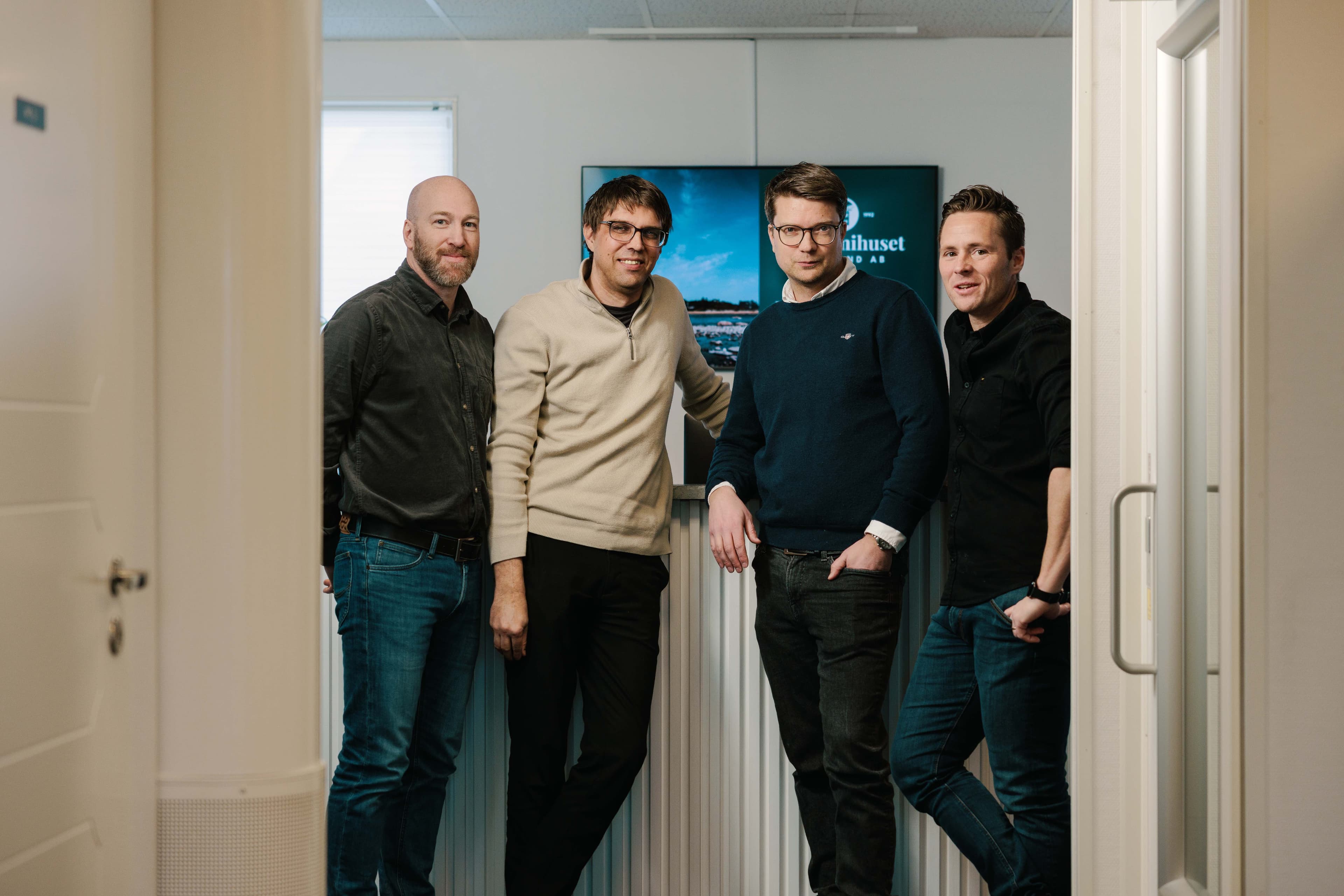 Four men stand together in a hallway, casually dressed, smiling at the camera with a sign in the background.