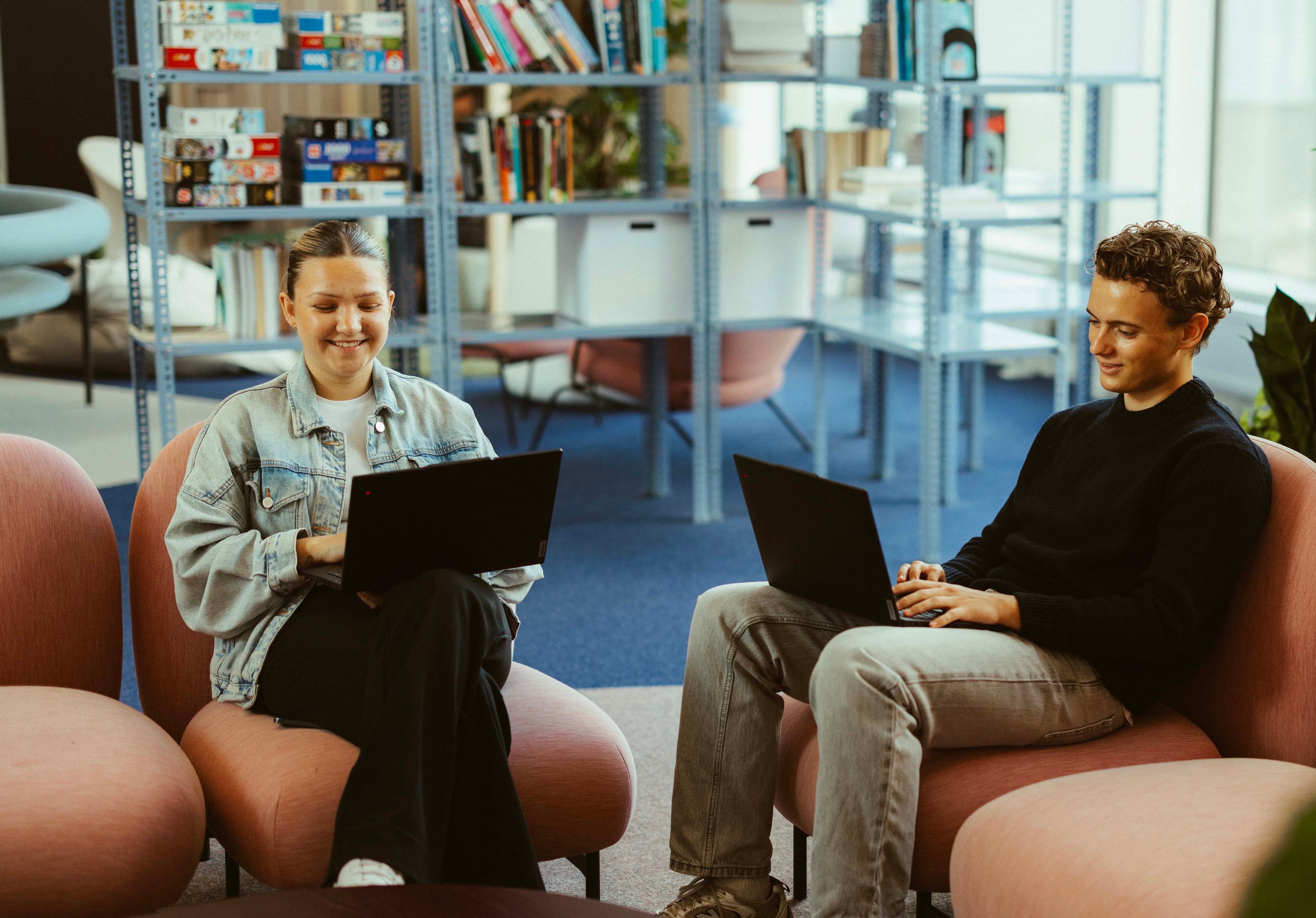 Two people sitting on pink chairs using laptops in a cozy library setting with bookshelves in the background.