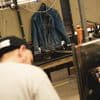 A denim jacket hangs on a pegboard in a workshop. A person with a baseball cap is blurred in the foreground, facing away.