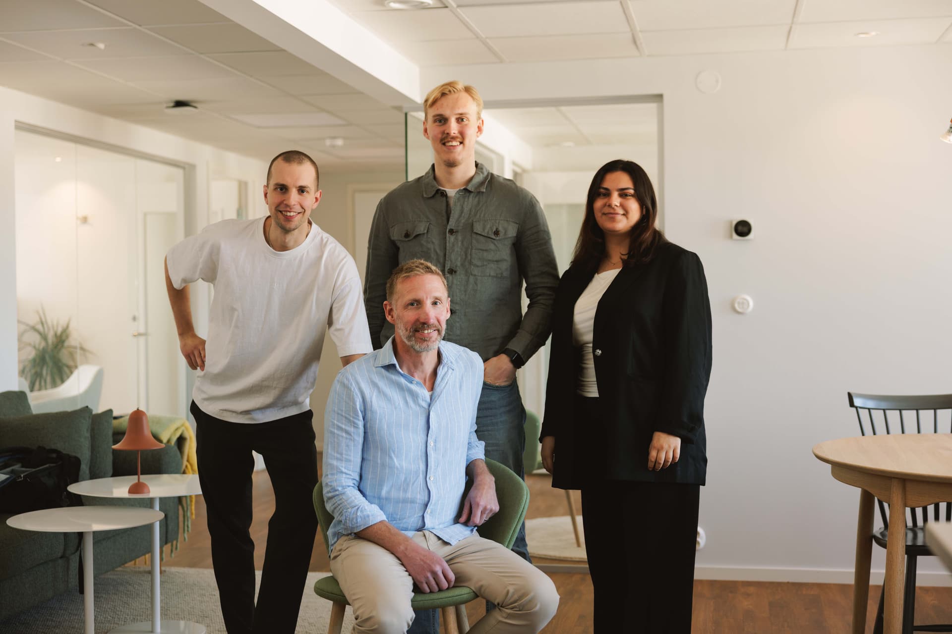 Four people posing in a modern office; one seated, three standing. They appear friendly and professional, with casual and business attire.