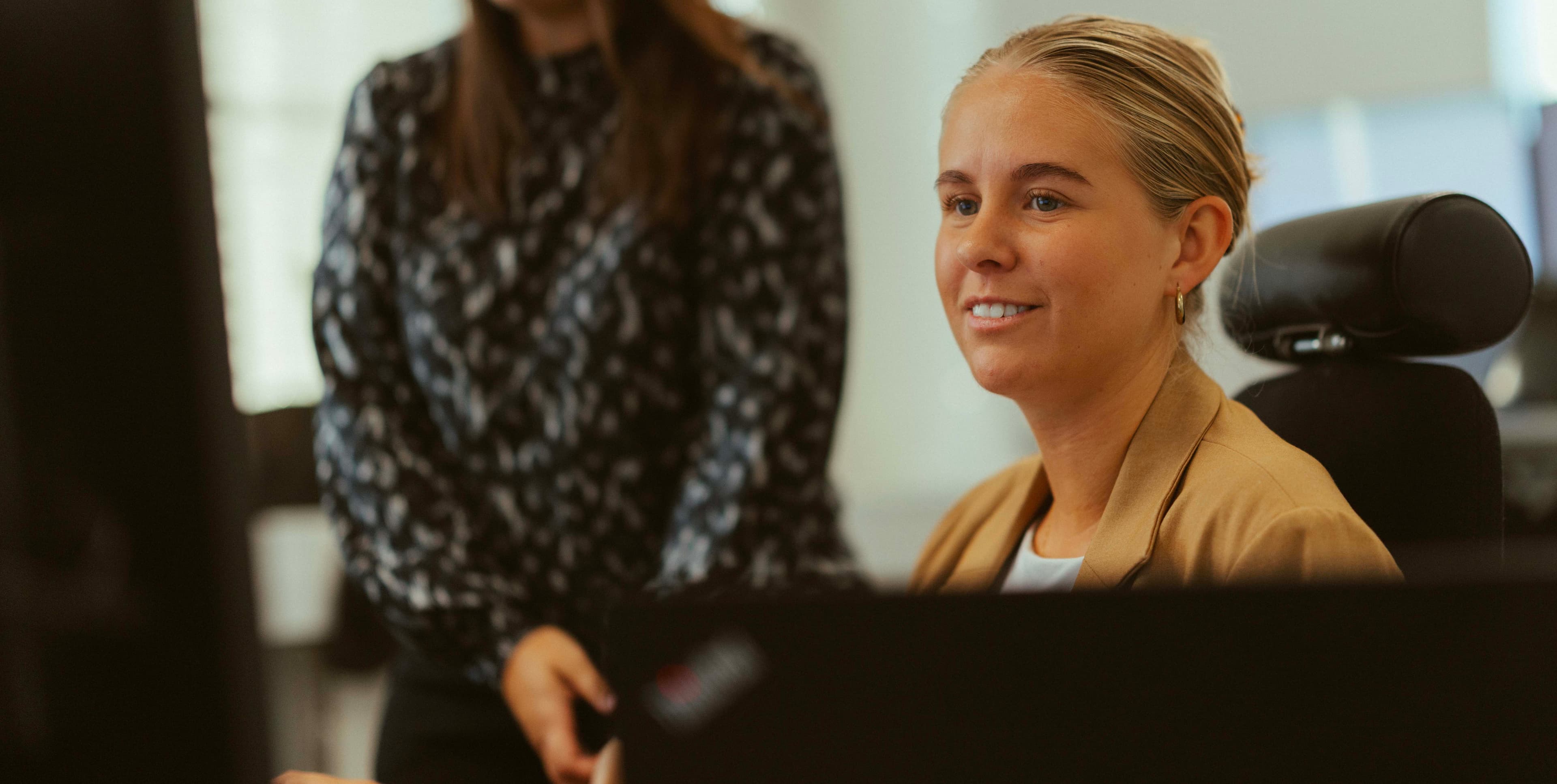 Two women working at a desk, one seated and smiling, the other standing and pointing at a computer screen.