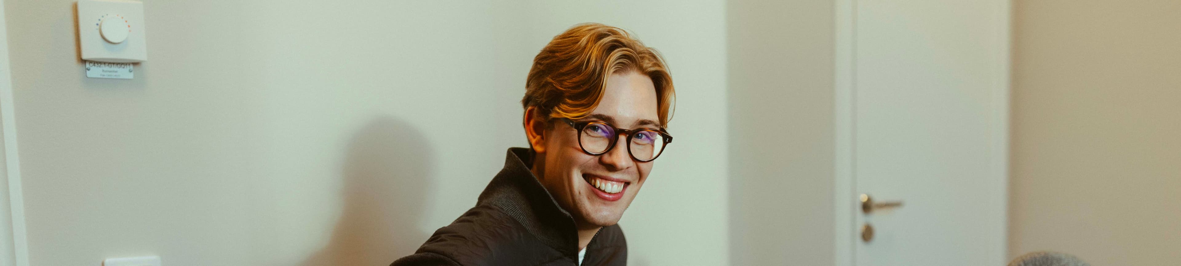 A smiling person with glasses and light brown hair, wearing a dark jacket, sits in a room with a thermostat on the wall.