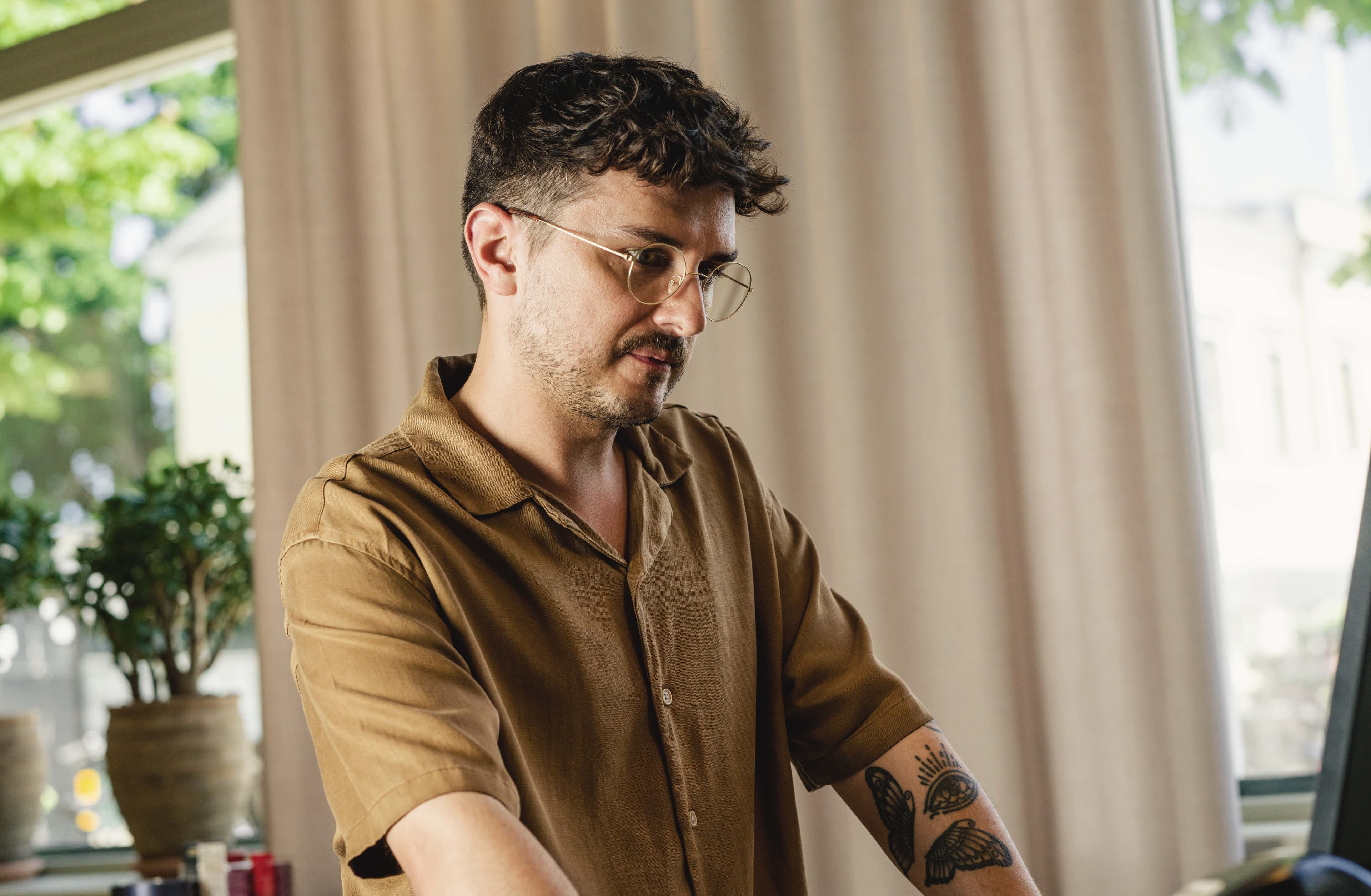 A man with glasses and a brown shirt works on a computer, with a tattoo visible on his arm and a potted plant in the background.