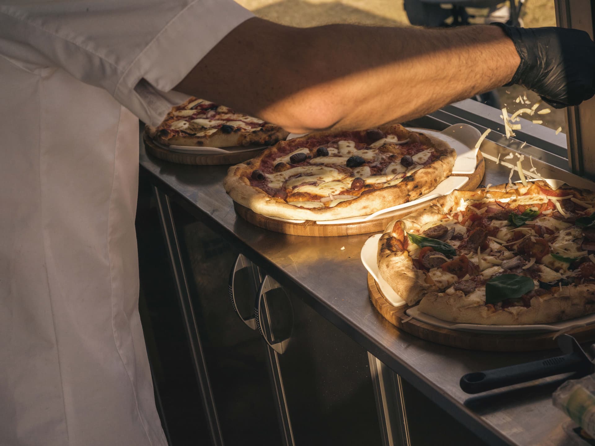 A person in a white coat is preparing pizzas with toppings on a wooden board, with shredded cheese being sprinkled on one pizza.